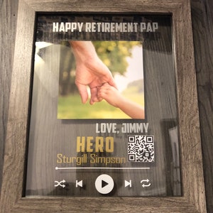 May include: A framed photo with a picture of two hands holding each other. The frame has a gray wood finish and the text "HAPPY RETIREMENT PAP LOVE, JIMMY HERO STURGILL SIMPSON" is printed on the glass. There is a QR code and a play button on the frame.