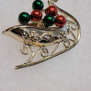 May include: A gold-tone metal Christmas sleigh pin with red and green glass beads. The sleigh is decorated with a holly leaf design.