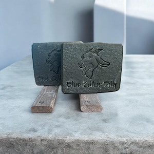 May include: Two dark green bars of soap with a goat head design and the text "Blue Collar Billy" on the bottom. The soap bars are sitting on wooden stands.