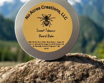 May include: A silver tin with a yellow label that reads "No Acres Creations, LLC" and "Sweet Tobacco Beard Balm". The label also includes a small black bee illustration and a list of ingredients.