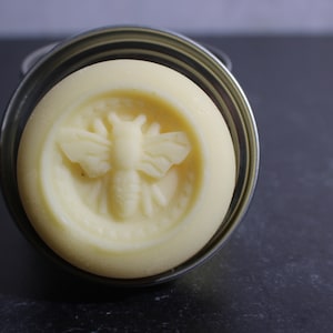 Puede incluir: Un bálsamo redondo, blanco y sólido con un diseño de abeja en relieve en la parte superior. El bálsamo está en una lata de plata.
