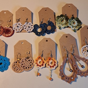 May include: A collection of handmade crochet earrings in various colors, including brown, white, blue, green, and orange. The earrings feature floral and circular designs, displayed on brown tags. Some earrings have beaded accents and dangle from silver or gold-colored hooks.