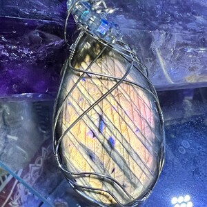 May include: A silver wire wrapped pendant featuring a large iridescent labradorite gemstone with a striated pattern. The pendant is displayed on a glass surface.