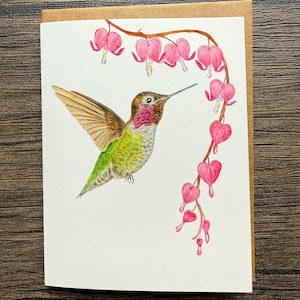 May include: A watercolor greeting card featuring a hummingbird in flight, with wings spread. The bird has green, red, and brown feathers. Pink bleeding heart flowers hang from a branch above. The card is on a white background with a brown envelope.