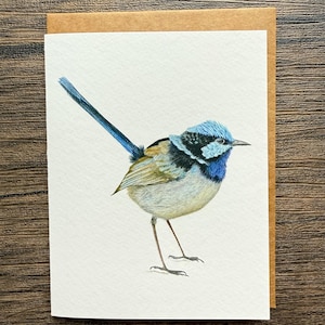 May include: A watercolor greeting card featuring a detailed illustration of a small bird with blue, black, and tan feathers. The bird is standing on thin legs against a white background, with a brown card backing.
