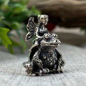 Könnte beinhalten: Ein silberner Anhänger mit einer Fee, die auf einem Frosch reitet. Die Fee hat Flügel und hält eine Blume. Der Frosch sitzt auf einem Sockel.