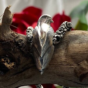 Peut inclure: Un bracelet en argent avec un charme en forme d'oiseau, présentant un design d'oiseau détaillé. Le bracelet est composé d'une chaîne avec une surface texturée. L'oiseau est perché sur une branche.