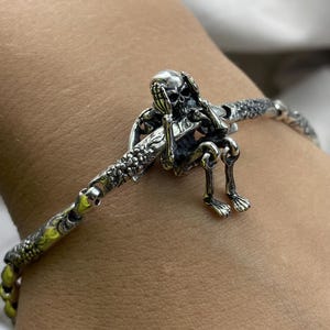 Peut inclure: Un bracelet en argent avec un charme de crâne détaillé. Le crâne se couvre les yeux avec ses mains. Le bracelet est fait d'une chaîne avec un design texturé.