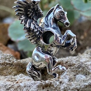 Peut inclure: Un charme en argent en forme de cheval ailé avec un design détaillé. Le cheval se tient sur ses pattes arrière, les ailes déployées. Le charme a un trou au centre pour l'attacher à un bracelet ou un collier.