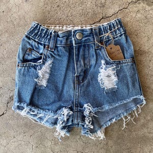 Puede incluir: Pantalones cortos vaqueros azules desgastados con bordes deshilachados y detalles rasgados. Los pantalones cortos tienen cierre de botón, bolsillos y una etiqueta de Baby Gap. Una prenda casual y elegante para niños.