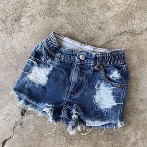 Puede incluir: Shorts vaqueros azules con detalles desgastados, incluyendo secciones rasgadas y bordes deshilachados. Los shorts tienen cierre de botón y cintura elástica. Un estilo casual para el uso diario.