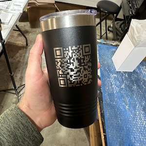 Può includere: Un bicchiere in acciaio inossidabile nero con un coperchio argentato e un codice QR inciso a laser. Il bicchiere è tenuto da una mano.