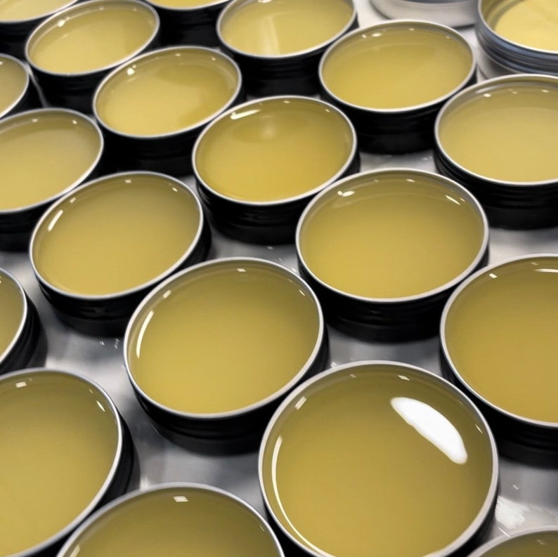 May include: A close-up view of multiple small round containers filled with a light yellow balm. The containers are made of metal and have a black lid.