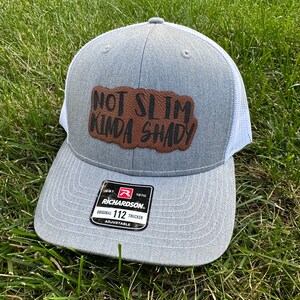 Puede incluir: Un gorro de camionero gris y blanco con un parche de cuero marrón que dice "Not Slim Kinda Shady". El gorro es ajustable y tiene una etiqueta Richardson 112.
