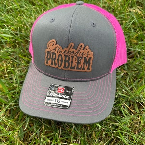 Puede incluir: Gorra de camionero gris y rosa con un parche de cuero marrón que dice "Somebody's Problem". La gorra es ajustable y tiene una etiqueta Richardson 112.