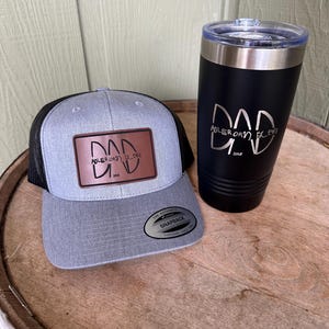 Może przedstawiać: Szara i czarna czapka typu trucker z brązową skórzaną naszywką z napisem "DAD" i "ADLER ORV, ELEK" pod logo. Czarny kubek ze srebrną obręczą i pokrywką ma to samo logo "DAD". Czapka i kubek są prezentowane na drewnianej powierzchni.