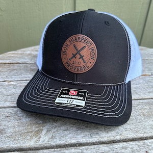 Könnte beinhalten: Schwarze und weiße Trucker-Kappe mit einem braunen Leder-Patch, auf dem "Iron Sharpens Iron 27:17 Proverbs" steht. Die Kappe ist verstellbar und hat ein Richardson-Logo.