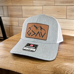 Könnte beinhalten: Graue und weiße Trucker-Kappe mit einem braunen Leder-Patch, der eine Bergkette und den Text "G>A>V" zeigt. Der Patch ist auf der Vorderseite der Kappe aufgenäht. Die Kappe hat einen verstellbaren Riemen mit einem schwarzen Etikett, auf dem "Richardson 112 Original Trucker Adjustable" steht.