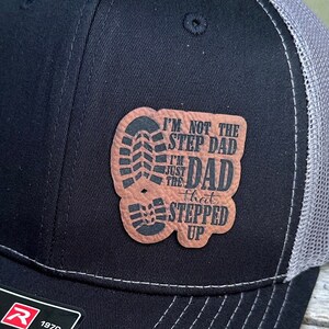 May include: Black trucker hat with a gray mesh back and a leather patch. The patch features a boot print and the text: "I'M NOT THE STEP DAD I'M JUST THE DAD that STEPPED UP."