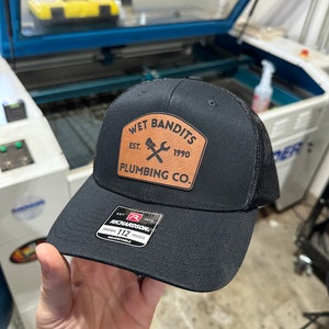 Może przedstawiać: Czarna czapka trucker z brązową skórzaną naszywką z napisem "Wet Bandits Est. 1990 Plumbing Co." i skrzyżowanym kluczem i rurą.