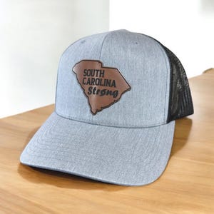 May include: A gray and black trucker hat with a brown leather patch featuring the outline of South Carolina and the text "South Carolina Strong".