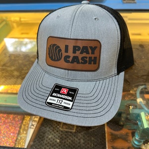 May include: A grey and black trucker hat with a brown leather patch that says "I PAY CASH". The hat has a black mesh back and a Richardson 112 adjustable label.