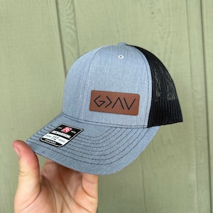 Könnte beinhalten: Eine graue und schwarze Trucker-Kappe mit einem braunen Leder-Patch, auf dem "G>N" steht.