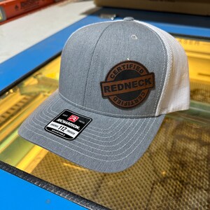 May include: Gray and white trucker hat with a curved brim. The hat features a leather patch with the words "CERTIFIED REDNECK" in a circular design. The hat has an adjustable strap and a Richardson tag.