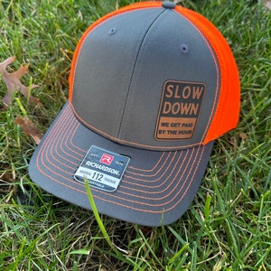 Può includere: Un cappello da camionista grigio e arancione con una toppa marrone che recita "Slow Down We Get Paid By The Hour". Il cappello ha un'etichetta Richardson 112 trucker.