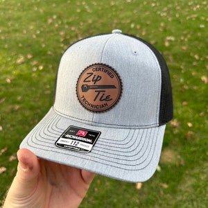 May include: A grey and black trucker hat with a brown leather patch that says "Certified Zip Tie Technician". The patch has a stylised image of a zip tie and the text "Original". The hat is a Richardson 112 Trucker.