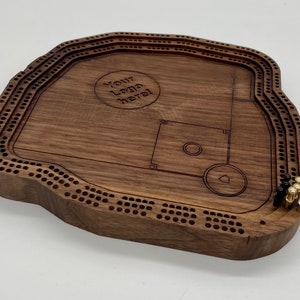 Op de afbeelding: Een houten cribbage-bord in de vorm van een honkbalveld met een aangepast logo in het midden. Het bord heeft een donkerbruine kleur en beschikt over een honkbalveld met een homeplate en een werpersheuvel. Het bord heeft een rij gaten rond de omtrek en een rij gaten rond de binnenkant van het honkbalveld. Er zijn vier zwarte, gouden en zilveren pinnen op het bord.