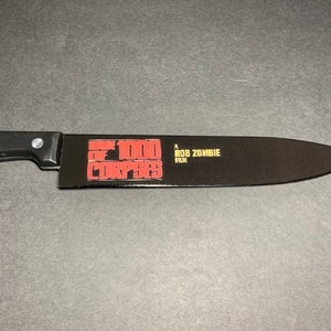 Puede incluir: Un cuchillo de cocina negro con mango negro. La hoja tiene un gr&aacute;fico rojo y blanco que dice "House of 1000 Corpses" y "A Rob Zombie Film".