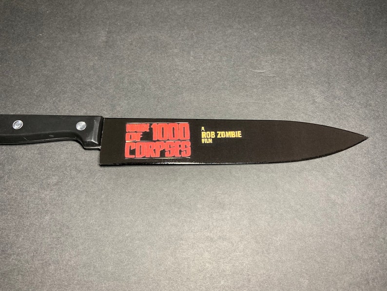Puede incluir: Un cuchillo de cocina negro con mango negro. La hoja tiene un gr&aacute;fico rojo y blanco que dice "House of 1000 Corpses" y "A Rob Zombie Film".