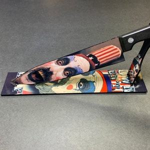 Puede incluir: Un cuchillo de cocina con mango negro con una imagen impresa de la cara de un payaso con un sombrero rojo, blanco y azul. El cuchillo se muestra en un soporte con una imagen impresa de la cara de un payaso y el texto "We All Float Down Here".