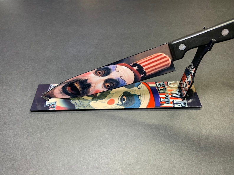 Puede incluir: Un cuchillo de cocina con mango negro con una imagen impresa de la cara de un payaso con un sombrero rojo, blanco y azul. El cuchillo se muestra en un soporte con una imagen impresa de la cara de un payaso y el texto "We All Float Down Here".