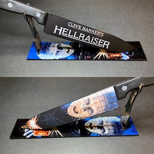 May include: A black knife with a silver handle and a black stand. The knife has the words "Clive Barker's Hellraiser" printed on it. The stand is decorated with a graphic of a character from the Hellraiser movie.