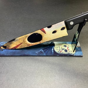 May include: A black-handled kitchen knife with a printed image of a hockey mask on the blade. The knife is displayed on a stand with a printed image of a hockey mask.