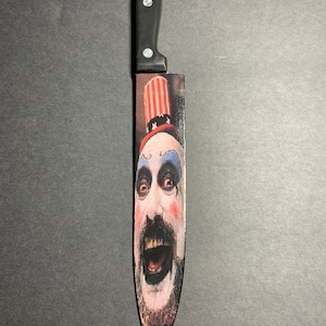 Puede incluir: Un cuchillo de cocina con mango negro y una imagen impresa de un payaso con un sombrero rojo, blanco y azul. El payaso tiene la cara blanca con ojos negros, un bigote negro y la boca abierta de par en par.