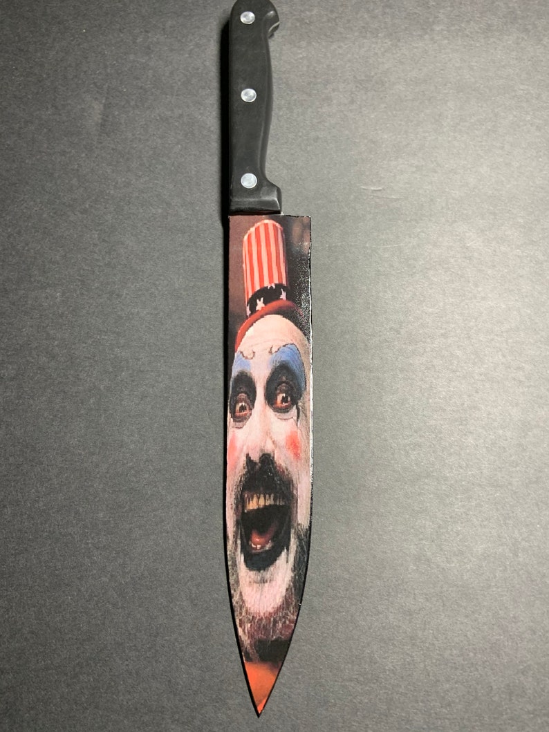 Puede incluir: Un cuchillo de cocina con mango negro y una imagen impresa de un payaso con un sombrero rojo, blanco y azul. El payaso tiene la cara blanca con ojos negros, un bigote negro y la boca abierta de par en par.