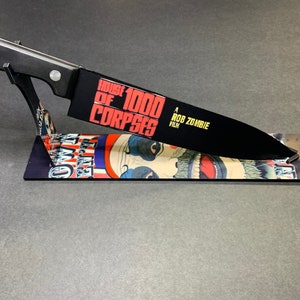 Puede incluir: Un cuchillo de cocina negro con mango negro y un gr&aacute;fico blanco y rojo en la hoja que dice "House of 1000 Corpses A Rob Zombie Film". El cuchillo est&aacute; en un soporte con un gr&aacute;fico de una cara de payaso.