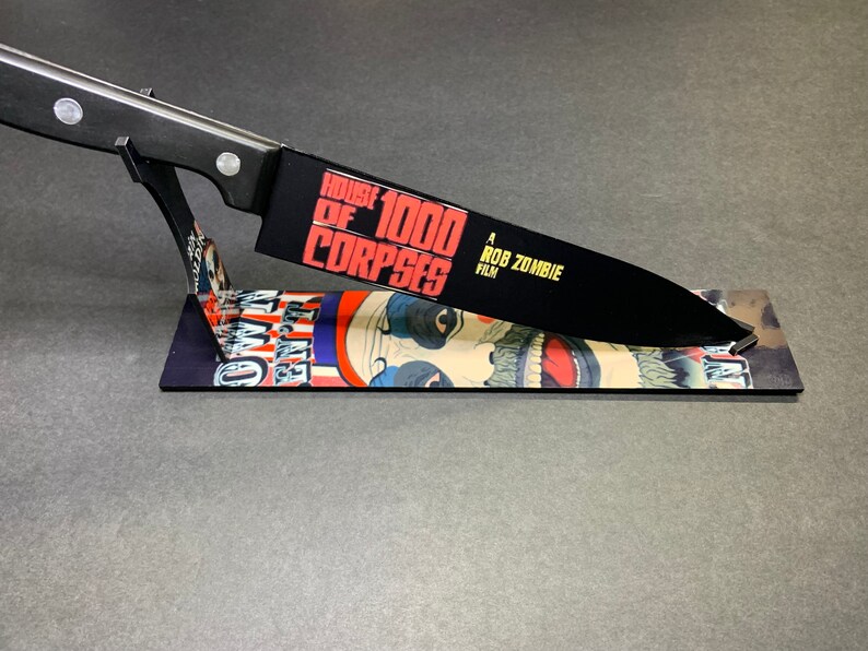 Puede incluir: Un cuchillo de cocina negro con mango negro y un gr&aacute;fico blanco y rojo en la hoja que dice "House of 1000 Corpses A Rob Zombie Film". El cuchillo est&aacute; en un soporte con un gr&aacute;fico de una cara de payaso.