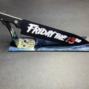 May include: A black knife with a white "Friday the 13th" inscription on the blade is displayed on a stand with a printed image of Jason Voorhees's hockey mask.