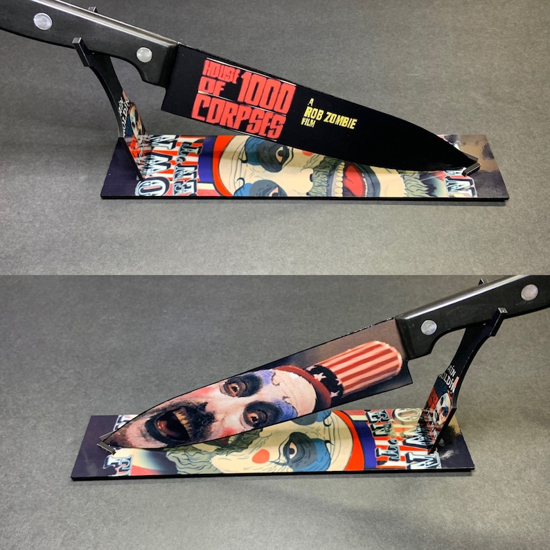 Puede incluir: Un cuchillo de cocina negro con mango blanco se muestra en un soporte. El cuchillo est&aacute; decorado con un gr&aacute;fico de un personaje de la pel&iacute;cula "House of 1000 Corpses", una pel&iacute;cula de Rob Zombie.