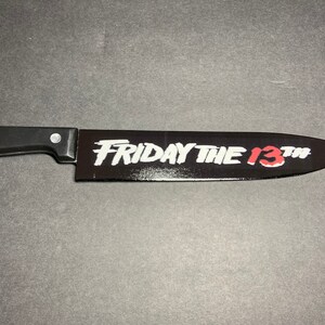 May include: A black plastic knife with a black handle. The blade has the words "FRIDAY THE 13TH" printed in white with the "13" in red.