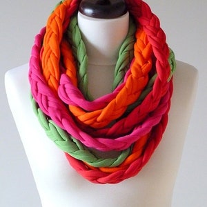 Op de afbeelding: Een gevlochten infinity sjaal in roze, oranje en groen. De sjaal is gemaakt van een zachte, rekbare stof en is perfect om een ​​kleuraccent toe te voegen aan elke outfit.