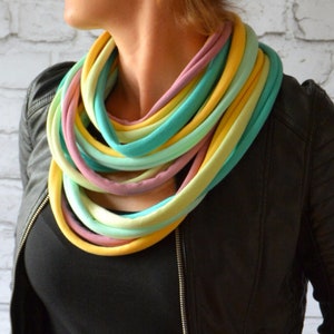 Op de afbeelding: Een veelkleurige stoffen ketting met een verloop van geel, groen, roze en paars. De ketting is gemaakt van een zachte, rekbare stof en wordt gedragen door een persoon met een zwart leren jack.