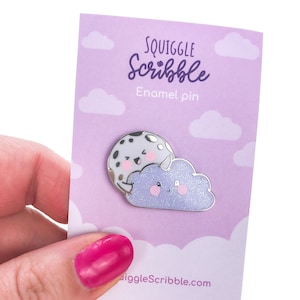 May include: A purple enamel pin featuring a cartoon cloud with a smiling face and a black and white moon with a smiling face. The pin is on a purple card with the text "Squiggle Scribble Enamel pin" and "gigglescribble.com".