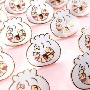 May include: A collection of gold-toned enamel pins shaped like a white steamed bun with a smiling shrimp inside. The shrimp is pink and orange with black eyes and a black mouth.