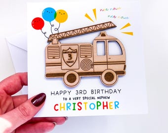 May include: A birthday card with a wooden fire engine design, a ladder, and three balloons. The card says "Happy 3rd Birthday to a very special nephew Christopher" in colourful letters. The card is held by a hand.