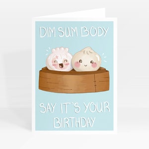 May include: A light blue greeting card with a cartoon illustration of two dim sum dumplings in a bamboo steamer. The text "Dim Sum Body Say It's Your Birthday" is printed on the card.